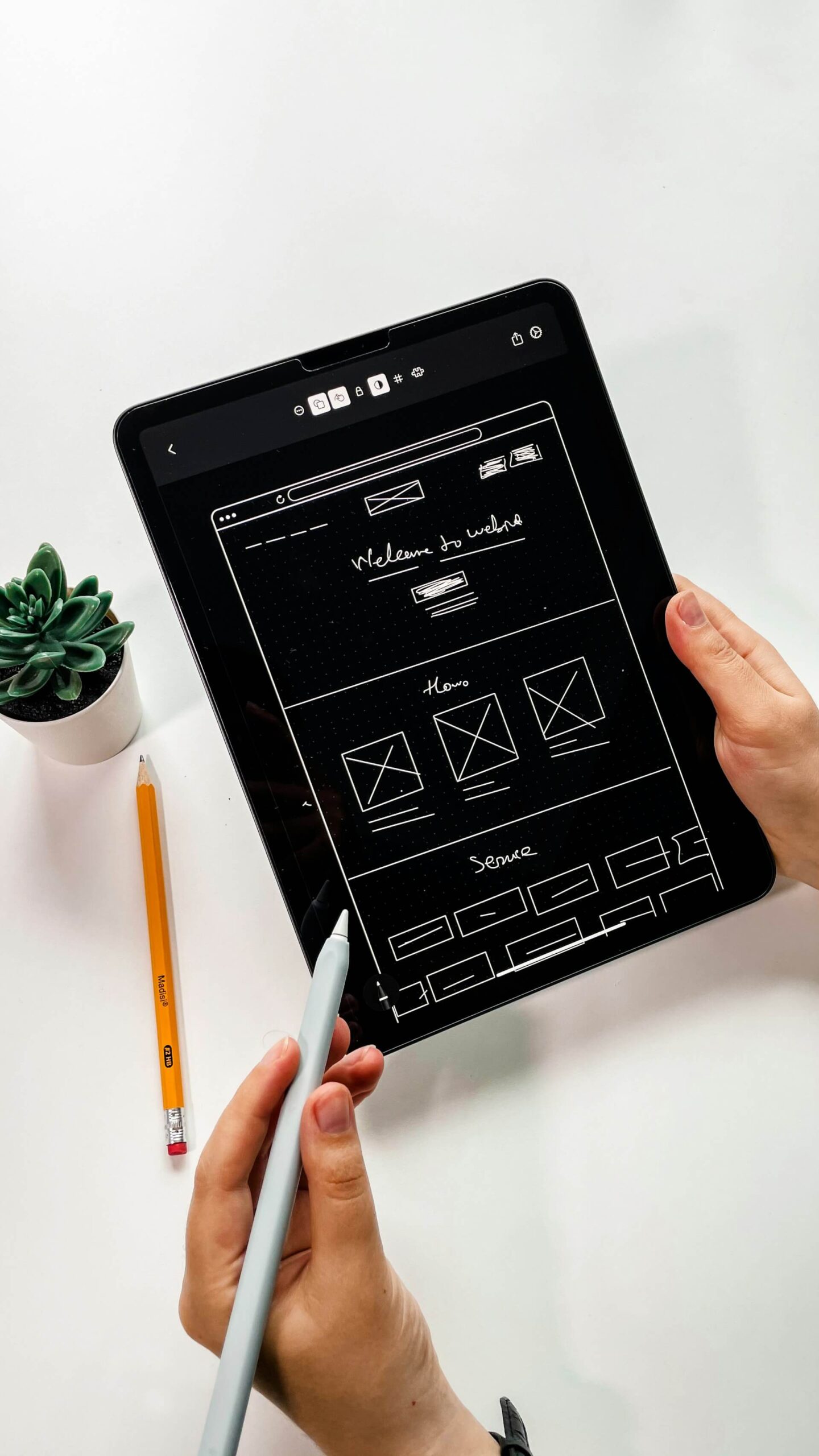Dos manos aguantando una tablet y el lapiz para la tablet con la estructura de la pagina web en la pantalla de la tablet. El fondo es blanco con un lapiz tradicional a la izquierda y una planta pequeña decorativa más arriba del lápiz.