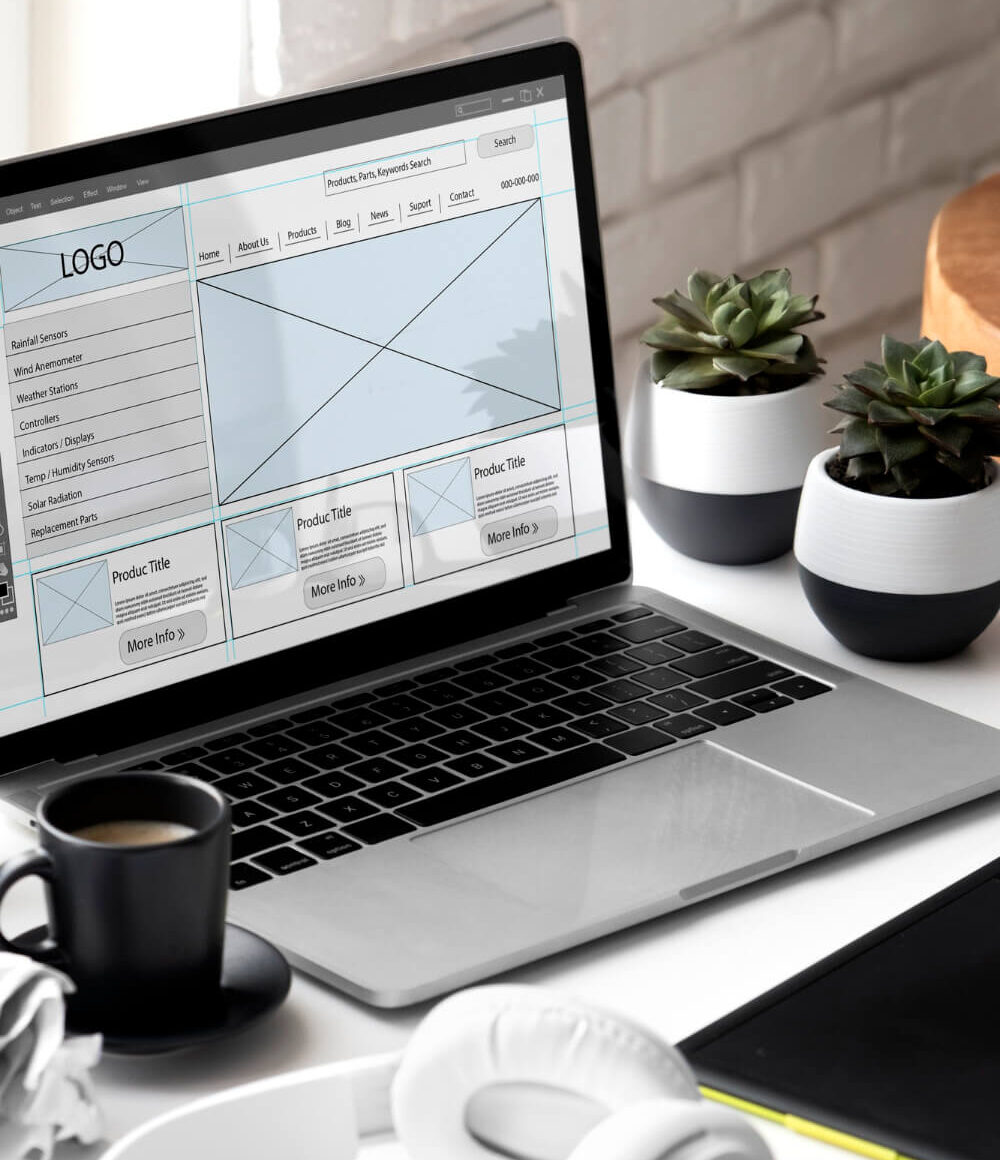 Estructura de una página web en la pantalla de una computadora. Hay una taza negra de café en el lado izquierdo de la computadora con unos audifonos blancos al frente de la taza. A la derecha hay dos plantas pequeñas decorativas.