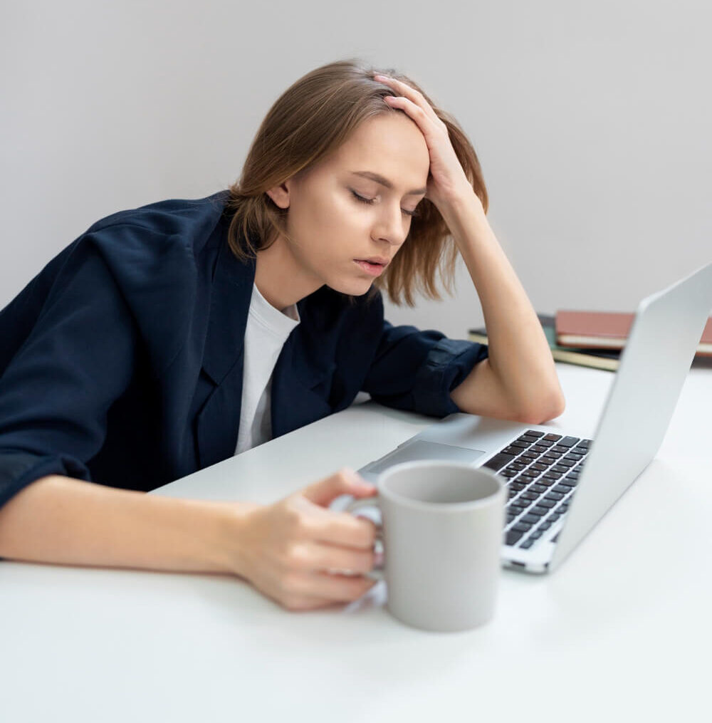 Mujer con camisa blanca y jacket azul oscuro sentada frente a la computadora con una taza a mano derecha. Parece estar abrumada y cansada. Tiene libros cerrados a la izquierda
