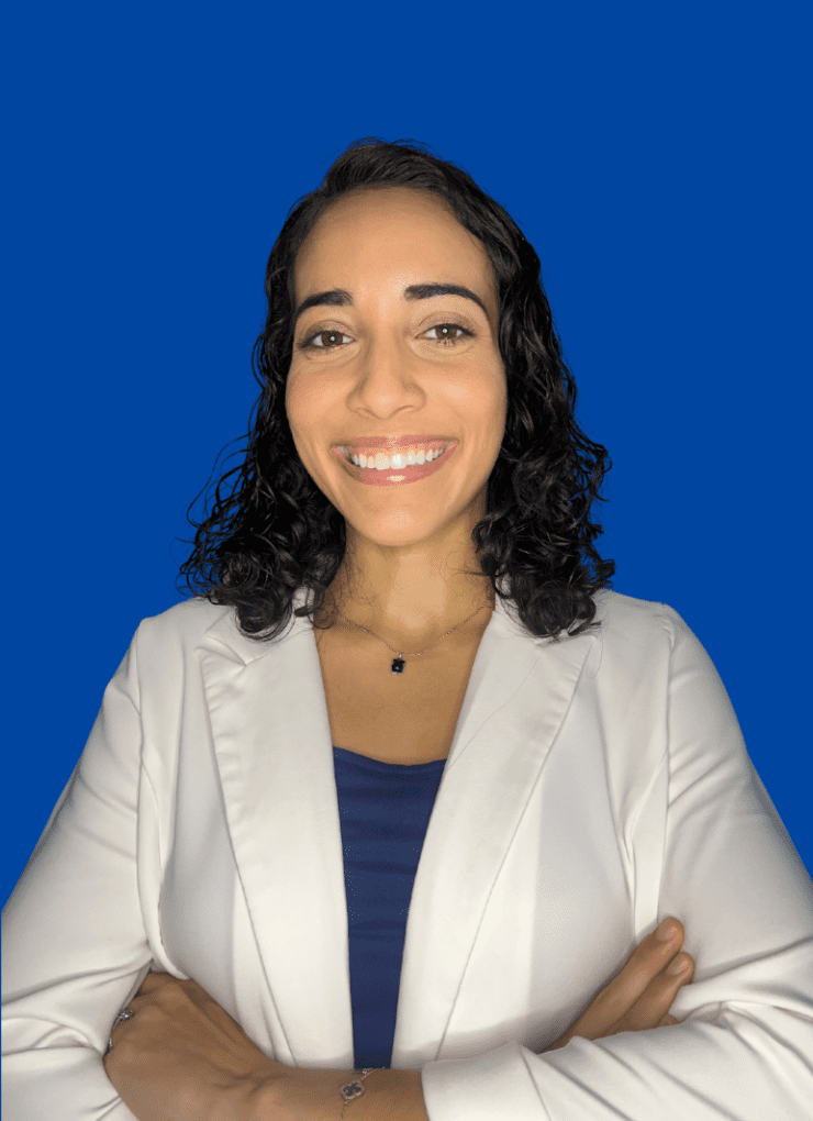 Monica Pérez con brazos cruzados, chaqueta blanca y blusa azul con un collar negro sonriendo hacia la cámara con un fondo azul.